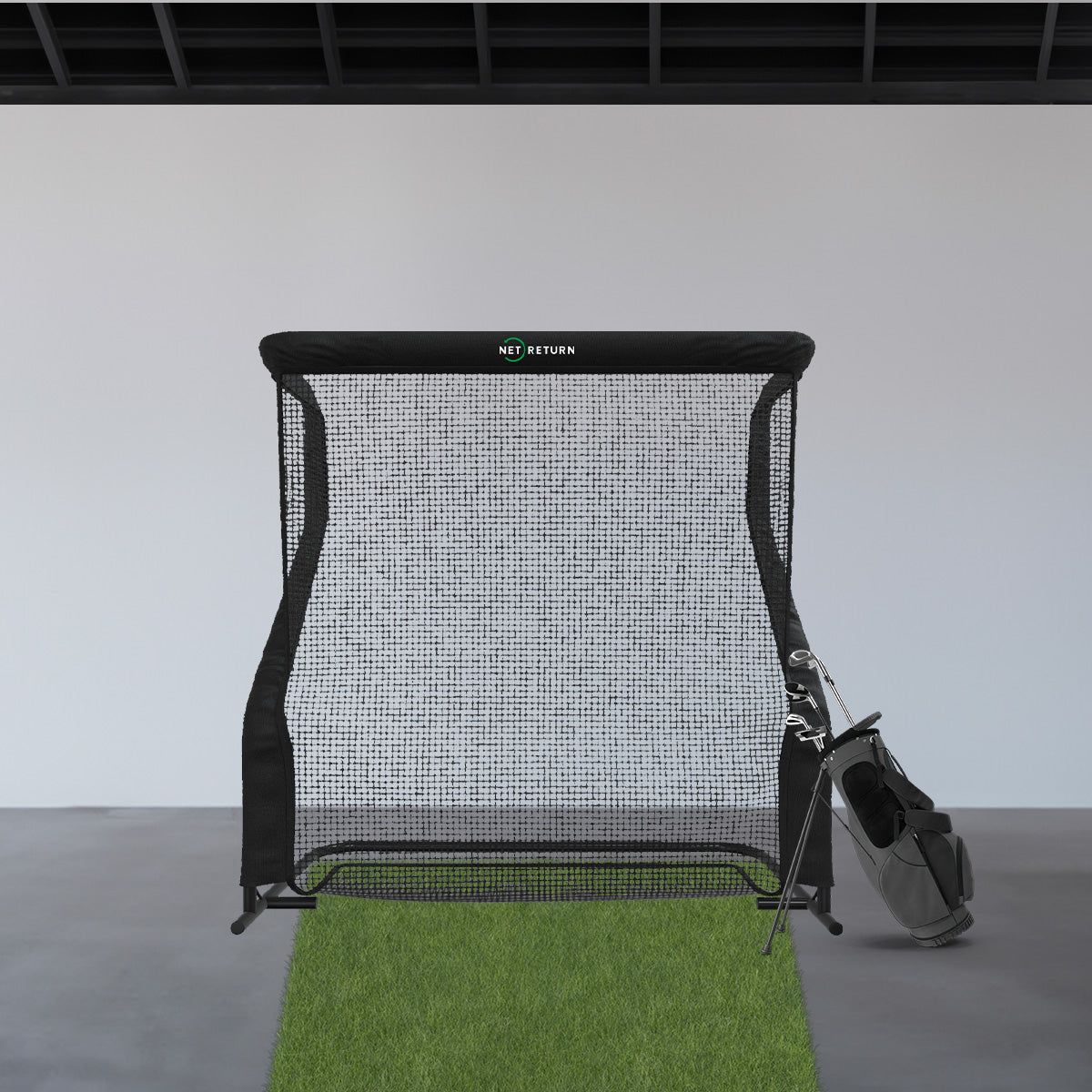 Golf net with green turf and a black golf bag holding clubs beside it, set indoors on a concrete floor.